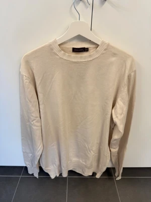 Beige rundhalsad tröja från Oscar Jacobsson, stl XL - Klassisk beige tröja från Oscar Jacobsson med rund halsringning och ribbade muddar vid ärmslut och nederkant. Enkel och stilren design som passar till det mesta. Tillverkad i mjukt material för en skön känsla.