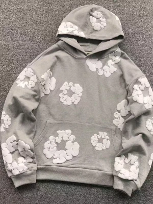 Denim tears hoodie med vita blommor - Säljer en grå hoodie med stora vita blommor i ett broderat mönster. Hoodien har en klassisk känguruficka framtill och en rymlig huva. Perfekt för dig som gillar en chill och unik stil.