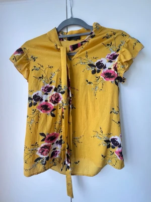 Gul blommig blus med knytband SHEIN - Gul blus från SHEIN med blommigt mönster i rosa, lila och svart. Blusen har korta ärmar med volangdetaljer och ett knytband vid halsen.