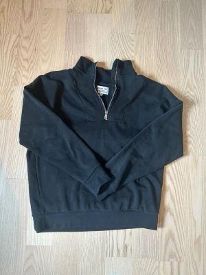 Svart halfzip sweatshirt Denim Day - Svart sweatshirt från Denim Day med halv dragkedja och hög krage. Tröjan har långa ärmar, ribbade muddar och är gjord i mjukt bomullsmaterial. Enkel och clean design utan tryck eller mönster, perfekt till jeans eller joggers.
