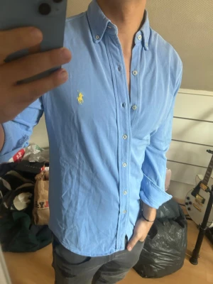 Blå skjorta från Polo Ralph Lauren - Säljer en klassisk blå skjorta från Polo Ralph Lauren med button-down krage och broderad gul logga på bröstet. Skjortan har långärmad design, knappar framtill och är tillverkad i mjuk bomull. Perfekt för dig som gillar stilrena plagg med ikonisk branding.