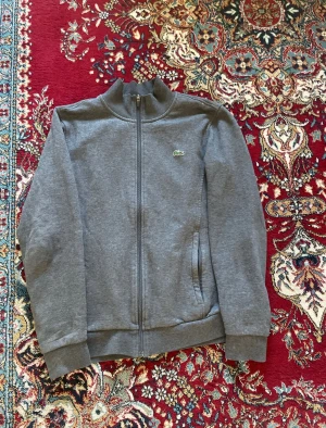 Grå ziptröja från Lacoste Sport - Säljer en stilren grå ziptröja från Lacoste Sport med hög krage och dragkedja framtill. Tröjan har två fickor och den klassiska gröna krokodilloggan på bröstet. Perfekt till chill eller streetwear. Mjuk bomullsblandning för skön känsla.