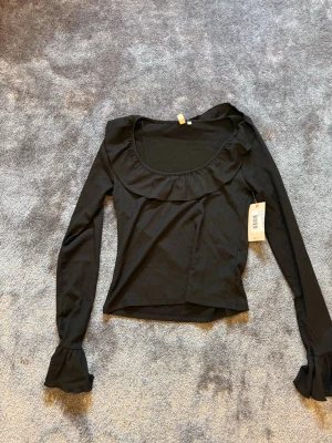 Svart långärmad topp med volang - Trendig svart topp med lång ärm och bred volangkant runt den djupa, rundade ringningen. Ärmsluten har också volangdetaljer som ger en extra touch. Perfekt att styla med jeans eller kjol för en cool look.