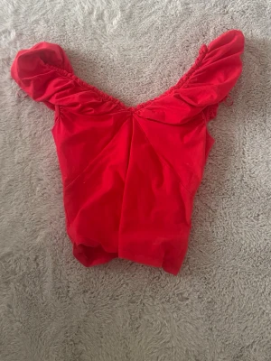 Röd offshoulder topp med volang - Snygg röd topp med offshoulder-design och volangdetaljer på axlarna. Perfekt för dig som vill ha en trendig och färgstark look. Materialet känns mjukt och stretchigt, vilket gör den bekväm att bära. Passformen är tight och axelbanden är breda.