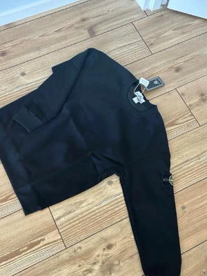 Svart sweatshirt från Stone Island - Snygg svart sweatshirt från Stone Island med klassisk rund hals och ribbade muddar. Tröjan har den ikoniska logotypen på ärmen och är tillverkad i mjukt bomullsmaterial. Perfekt för en clean och stilren look. OBS Tröjans storlek är M men passar som S-XS