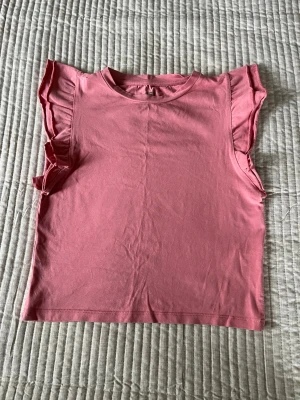 Rosa topp med volangärmar - Söt rosa topp i mjuk bomull med volangdetaljer vid ärmarna. Modellen är ärmlös med en rak passform och rund halsringning. Perfekt för dig som vill ha en enkel men ändå trendig topp till sommaren.