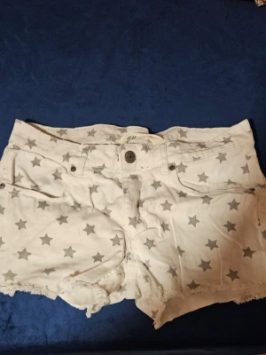Vita shorts med stjärnor från H&M - Snygga vita shorts från H&M med gråa stjärnor över hela tyget. Modellen har fransig kant nertill, klassisk femficksdesign och knappgylf. Perfekta för sommaren och ger en cool vibe till din outfit.