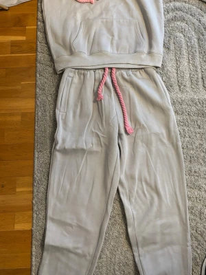 Syna world set - Mysigt loungewear-set i ljusgrått med hoodie och byxor. Setet har rosa dragsko och broderad text 'Imagine Me' på byxbenet. Tillverkat i mjukt material, perfekt för chill dagar hemma. Praktisk magficka på hoodien och sidofickor på byxorna.