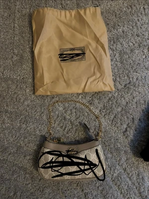 Dam väska - Snygg beige kuvertväska   med klassiskt GG-mönster och guldfärgad kedja som axelrem. Väskan har detaljer i ljusbrunt läder och dragkedja upptill. Perfekt accessoar för att lyfta din outfit.