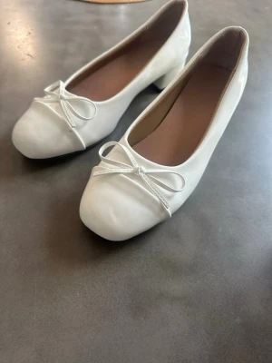 Vita ballerinaskor med rosett - Snygga vita ballerinaskor med låg klack och söt rosett framtill. Skorna har rundad tå och är gjorda i ett glansigt material som ger en fräsch look. Perfekta för dig som gillar klassisk och stilren design.