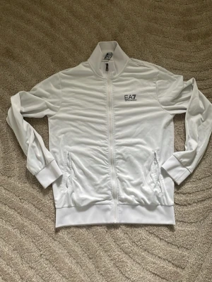 Vit ziptröja från EA7 Emporio Armani - Snygg vit ziptröja från EA7 Emporio Armani med hög krage och dragkedja framtill. Tröjan har två fickor med dragkedja och diskret logga på bröstet. Har haft den ganska länge men den har inga nedsättningar som syns.