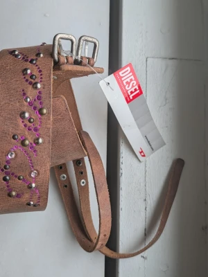 Brunt skärp med nitar från Diesel - Snyggt brunt skärp från Diesel i skinn med dubbla spännen. Skärpet har dekorativa nitar och lila stenar i ett mönster längs hela bandet. Perfekt accessoar för att lyfta din outfit med en cool detalj. 70 cm lång