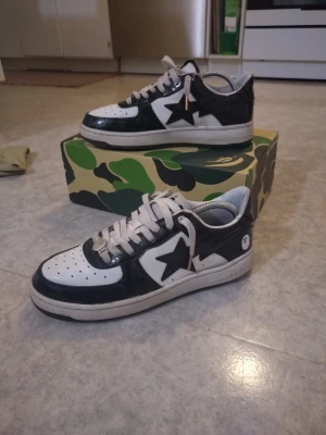 BAPE STA svartvita sneakers - Säljer ett par BAPE STA sneakers i svart och vitt med den ikoniska stjärnan på sidan. Skorna har låga skaft, perforerad tåbox och vita snören. Ovandelen är i blankt läder och sulan är vit med BAPE-logga på hälen. Kommer med originalkartong i camomönster.