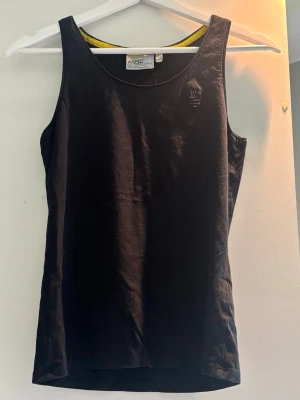 Svart linnetopp från WESC - Säljer en svart linnetopp från WESC med rund halsringning och diskret broderad logga på bröstet. Toppen är ärmlös och har en enkel, clean design med gul detalj på insidan av halsen. Perfekt för en avslappnad och stilren look.