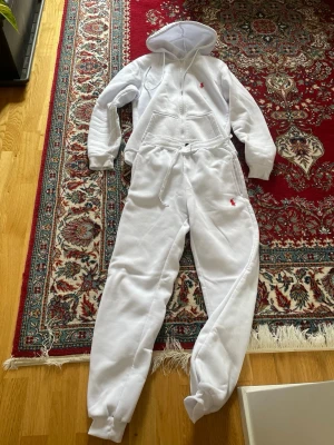 Vit jumpsuit med huva från Ralph Lauren - Säljer en vit jumpsuit från Ralph Lauren med dragkedja framtill, huva och fickor. Jumpsuiten har resår vid ärm- och benslut samt dragsko i midjan. Ikoniska röda Polo-loggan broderad på bröstet och benet. Perfekt för en avslappnad och sportig stil.