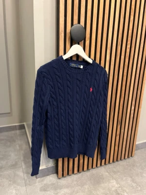 Kabelstickad kofta - Säljer en mörkblå kabelstickad tröja i storlek S. Tröjan är helt ny och oanvänd, lapparna sitter fortfarande kvar. Väldigt fin passform och klassisk modell med röd logga framtill. Säljes på grund av felköp. Perfekt skick utan några anmärkningar.
