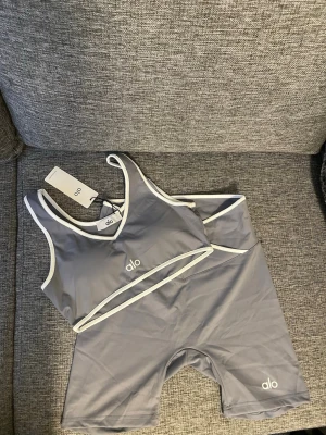 Grått sportigt set från Alo - Snyggt och sportigt set från Alo i grått med vita kanter. Setet består av en crop top-bh med breda axelband och matchande cykelbyxor, båda med Alo-logga framtill. Materialet är stretchigt och följsamt, perfekt för en aktiv livsstil.