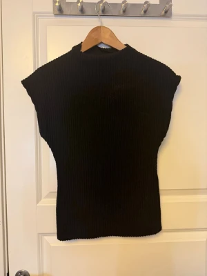 Svart ribbad ärmlös topp - Snygg svart ribbad topp med hög hals och ärmlös design. Perfekt att styla med jeans eller kjol för en clean och trendig look. Materialet är stretchigt och följsamt för en bekväm passform.