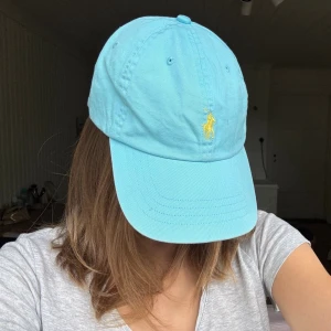 Ljusblå keps från Polo Ralph Lauren - Snygg ljusblå keps från Polo Ralph Lauren med den klassiska gula loggan broderad framtill. Kepsen har böjd skärm och justerbar rem bak. Tillverkad i bomull för en skön och avslappnad look. 