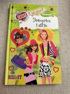 Drömprins i sikte av Anna Wellenberg - Färgglad ungdomsbok med hårda pärmar, illustrerad framsida i grönt, rosa och gult med två tecknade tjejer och hjärtan. Boken heter 'Drömprins i sikte' och är skriven av Anna Wellenberg. Perfekt för dig som gillar tjejkompisar, kärlek och vardagsdrama.