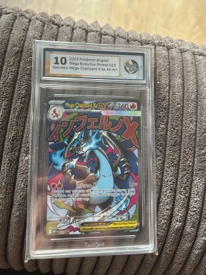 Mega Charizard X EX Alt Art Pokémonkort - Säljer ett graderingbedömt Mega Charizard X EX Alt Art Pokémonkort, engelskspråkig promo från 2025. Kortet har färgstark design med blå, svart och röd drake och är innesluten i skyddande plastkapsel från RaukCard.