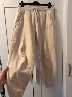 Beige mjukisbyxor med snöre - Säljer ett par beige mjukisbyxor med resår och dragsko i midjan. Byxorna har två sidofickor och ribbade muddar vid bensluten. Perfekta för chill dagar och riktigt sköna att ha på sig. Materialet känns mjukt och bekvämt.