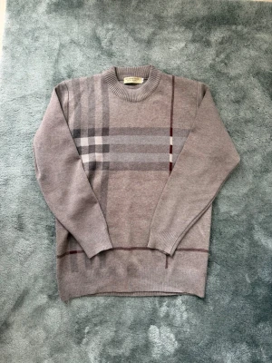 Stickad Burberry tröja - Snygg grå stickad tröja från Burberry med klassiskt rutmönster i olika nyanser av grått och vinrött. Tröjan har rund halsringning och ribbade muddar vid ärmslut och nederkant. Perfekt för dig som gillar stilrena och tidlösa plagg.