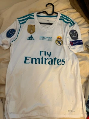 Real Madrid Ronaldo matchtröja 2018 - Säljer en vit Real Madrid matchtröja från Champions League-finalen 2018 med Ronaldo 7 på ryggen. Tröjan har blå detaljer, officiella märken och Climacool-material för bästa komfort. Fly Emirates-logga på bröstet och UEFA-patchar på ärmarna.