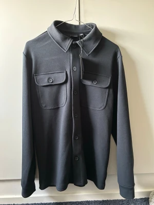 Mörkblå overshirt  - Stilren mörkblå skjortjacka med två stora bröstfickor med lock och knapp från                Only & Sons. Långa ärmar, klassisk krage och knäppning framtill. Materialet är stretchigt och sitter mer som storlek S. Aldrig använd prislapp kvar. Den är mörkare i verkligheten än va den ser ut på bilderna.