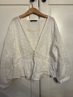 Vit broderad blus med knappar - Söt vit blus i broderad bomull med v-ringning och klädda knappar framtill. Blusen har lång ärm med resår vid ärmslut och ett lätt utsvängt avslut. Perfekt för en romantisk och bohemisk stil. Kort i modellen