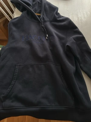 Mörkblå hoodie från Hackett London - Snygg mörkblå hoodie från Hackett London med broderad logga på bröstet. Klassisk känguruficka framtill och justerbar huva med dragsko. Perfekt för en chill och avslappnad stil. Mjuk bomullsblandning som känns skön mot huden.