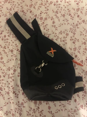 Svart ryggsäck från X-Over - Säljer en svart ryggsäck från X-Over med justerbar axelrem och reflexdetaljer. Väskan har ett lock med metallhake, tre metallöljetter och en orange dragflik. Tillverkad i slitstark textil, perfekt för dig som vill ha en smidig och cool väska.