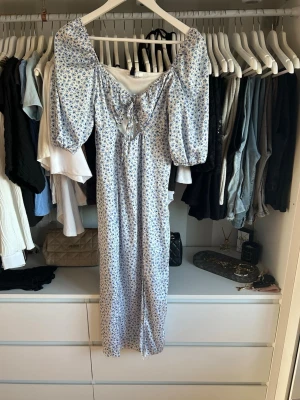 Blommig långklänning från H&M Divided - Supersöt långklänning från H&M Divided med blått blommönster på vit botten. Klänningen har puffärmar, snörning framtill och en romantisk v-ringning. Perfekt för sommarens alla tillfällen och tillverkad i ett lätt och skönt material.