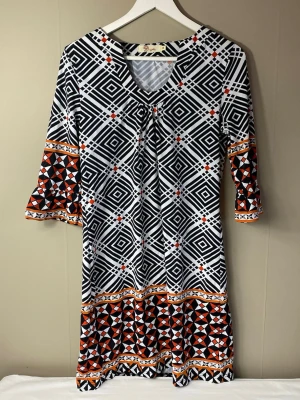 Mönstrad klänning med trekvartsärm - Snygg klänning från med grafiskt svartvitt rutmönster och inslag av orange och rött. Klänningen har trekvartsärm med volang och v-ringning. Materialet känns mjukt och stretchigt. Perfekt för dig som gillar färg och mönster!