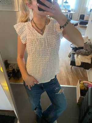 Vit blus med volangärmar - Söt vit blus med diskret rutigt mönster och volangdetaljer vid ärmarna. Blusen har v-ringning och är tillverkad i ett lätt, luftigt material som passar perfekt till jeans eller kjol. Kortärmad och något croppad modell för en trendig look.