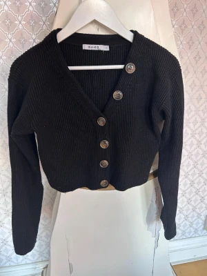 Svart ribbstickad kofta från NA-KD - Snygg svart ribbstickad kofta från NA-KD med v-ringning och fem stora knappar framtill. Kortare modell med långa ärmar och grovstickad struktur. Perfekt att slänga över en topp för en chill look.