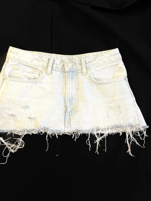 Ljusblå jeanskjol med fransig kant - Säljer en kort ljusblå jeanskjol med rå, fransig kant och slitna detaljer. Kjolen har klassisk femficksdesign, bälteshällor och knappgylf. Perfekt för dig som gillar en avslappnad och trendig stil. Stolek S 