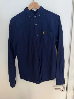 Mörkblå skjorta från Lyle & Scott - Klassisk mörkblå skjorta från Lyle & Scott med knappkrage och gul logga på bröstfickan. Skjortan har långa ärmar, knappar framtill och är tillverkad i mjuk bomull. Perfekt till jeans eller chinos för en clean look.