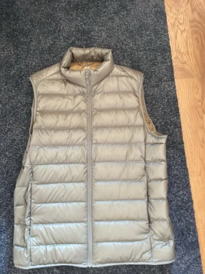 Beige dunväst från Uniqlo - Stilren beige dunväst från Uniqlo med hög krage och dragkedja framtill. Västen har en quiltad design och insidan är ljusgul. Perfekt för lager-på-lager och enkel att matcha med olika outfits.