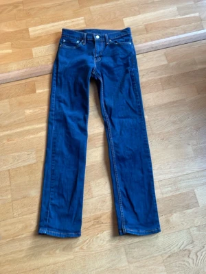 Blå raka jeans från Levi's - Säljer ett par klassiska blå jeans från Levi's med rak passform och fem fickor. Jeansen har silverfärgade nitar och knapp, samt den ikoniska läderlappen bak i midjan. Perfekta till vardags och passar till det mesta. Materialet är slitstark denim i bomull.