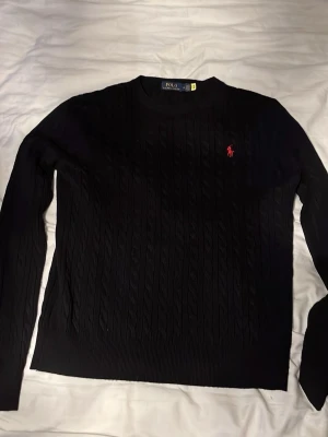 Stickad tröja Polo Ralph Lauren - Stickad svart tröja från Polo Ralph Lauren med rund halsringning och den klassiska röda logotypen broderad på bröstet. Långa ärmar och ribbade muddar. Perfekt för dig som gillar stilrena och tidlösa plagg.