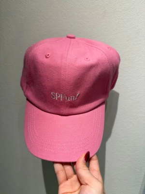 Rosa GLOWiD keps med broderad text Oanvänd - Snygg rosa keps i klassisk modell med böjd skärm och broderad vit text 'SPFun!' framtill. Kepsen har ventilationshål och justerbar rem bak. Perfekt accessoar för en trendig och avslappnad stil.