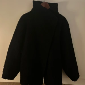 Svart teddyjacka med bälte - Svart teddyjacka med hög krage och bälte i midjan. Jackan har en oversized passform, lång ärm och grov textur. Perfekt för kalla dagar och ger en cool, modern look. Stängs med knappar och bälte för extra stilpoäng. Ordinarie pris: 600kr (många intresserade så säljer för endast 400kr) 