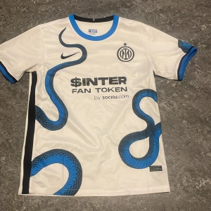 Inter Milan bortatröja Nike - Säljer en vit Inter Milan bortatröja från Nike med blå och svart orm-design, klubbmärke och sponsortryck på bröstet. Tröjan har korta ärmar, rund hals och blå detaljer vid ärmslut och krage. Perfekt för fotbollsfans som vill sticka ut.