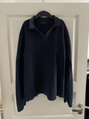 Mörkblå pikétröja från Riley - Klassisk mörkblå pikétröja från Riley med krage och v-ringad öppning utan knappar. Tröjan har lång ärm och är tillverkad i ett mjukt, stickat material som ger en avslappnad men ändå stilren look.