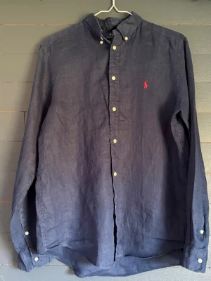 Mörkblå linneskjorta från Polo Ralph Lauren - Klassisk mörkblå skjorta i linne från Polo Ralph Lauren med vit knappstängning och röd broderad logga på bröstet. Skjortan har lång ärm och button-down krage, perfekt för en stilren och avslappnad look.