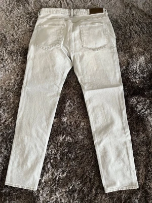 Ljusbeiga raka jeans - Säljer ett par ljusbeiga jeans med rak passform och klassisk femficksdesign. Jeansen har bälteshällor och knappgylf. Materialet är kraftig denim i bomull som ger en stilren och clean look. Perfekta för dig som gillar minimalistisk stil.