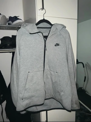 Grå zip hoodie från Nike - Säljer en grå zip hoodie från Nike med svart logga på bröstet. Hoodien har dragkedja hela vägen, två stora fickor fram och en skön huva. Tillverkad i mjukt material som passar perfekt till chill eller streetwear. Enkel och clean design med svarta detaljer.