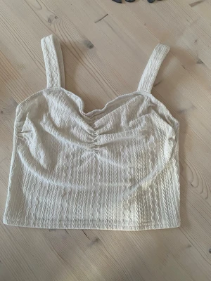 Beige croppad linnetopp med struktur - Säljer en vit croppad linnetopp med breda axelband och vågiga sömmar som ger en snygg struktur. Toppen har lätt rynkning vid bysten och är perfekt för varma dagar. Passar till både jeans och kjol för en trendig look. Köparen står för frakt.