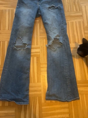Blå slitna bootcut jeans - Ett par blå bootcut jeans med stora slitningar över knäna och lätt utsvängda ben. Jeansen har klassisk femficksdesign och är gjorda i denim. Perfekta för en avslappnad och trendig look.